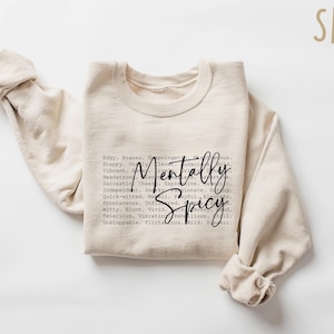 May include: A sand-colored sweatshirt with the words "Mentally Spicy" in a black script font. The sweatshirt has long sleeves and a crew neck. The text is surrounded by a list of adjectives.