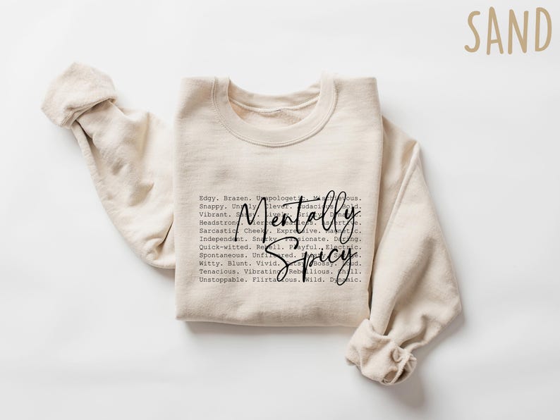 May include: A sand-colored sweatshirt with the words "Mentally Spicy" in a black script font. The sweatshirt has long sleeves and a crew neck. The text is surrounded by a list of adjectives.