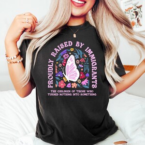 May include: Black t-shirt with the text "PROUDLY RAISED BY IMMIGRANTS" in a pink arch, featuring a butterfly and floral design. The shirt also includes the text "THE CHILDREN OF THOSE WHO TURNED NOTHING INTO SOMETHING".