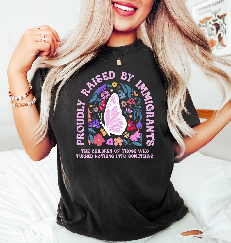 May include: Black t-shirt with the text "PROUDLY RAISED BY IMMIGRANTS" in a pink arch, featuring a butterfly and floral design. The shirt also includes the text "THE CHILDREN OF THOSE WHO TURNED NOTHING INTO SOMETHING".