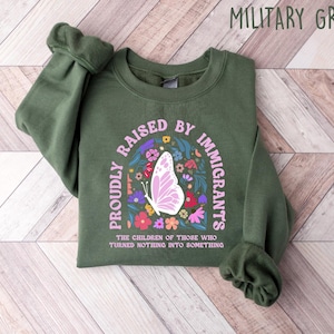 May include: Military green sweatshirt with the text "PROUDLY RAISED BY IMMIGRANTS" in a semi-circle around a butterfly and floral design. The text "THE CHILDREN OF THOSE WHO TURNED NOTHING INTO SOMETHING" is below the design.