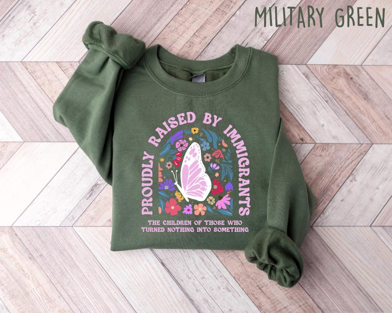 May include: Military green sweatshirt with the text "PROUDLY RAISED BY IMMIGRANTS" in a semi-circle around a butterfly and floral design. The text "THE CHILDREN OF THOSE WHO TURNED NOTHING INTO SOMETHING" is below the design.