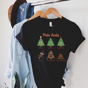 Puede incluir: Camiseta negra con un gr&aacute;fico humor&iacute;stico de "Pain Scale" con &aacute;rboles de Navidad de dibujos animados y texto. La escala va de "No Pain" a "Worst Pain Possible". La camiseta est&aacute; colgada en una percha de madera.