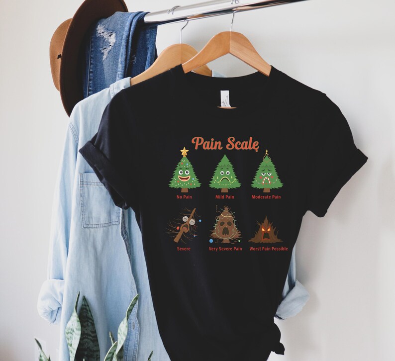 Puede incluir: Camiseta negra con un gr&aacute;fico humor&iacute;stico de "Pain Scale" con &aacute;rboles de Navidad de dibujos animados y texto. La escala va de "No Pain" a "Worst Pain Possible". La camiseta est&aacute; colgada en una percha de madera.