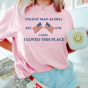 May include: A light pink t-shirt with the text "I'M JUST MAD AS HELL EST. 1776 CAUSE I LOVED THIS PLACE" and two American flags. The shirt is paired with light blue shorts. A white mug with a yellow smiley face is held in the hand.