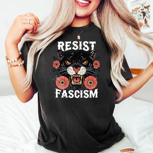 May include: Black t-shirt with the words "RESIST FASCISM" and a panther graphic. The panther has yellow eyes and is surrounded by red and orange flowers. The shirt is a dark color.