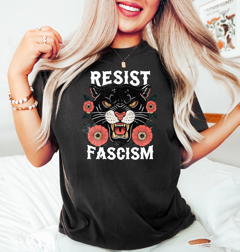 May include: Black t-shirt with the words "RESIST FASCISM" and a panther graphic. The panther has yellow eyes and is surrounded by red and orange flowers. The shirt is a dark color.