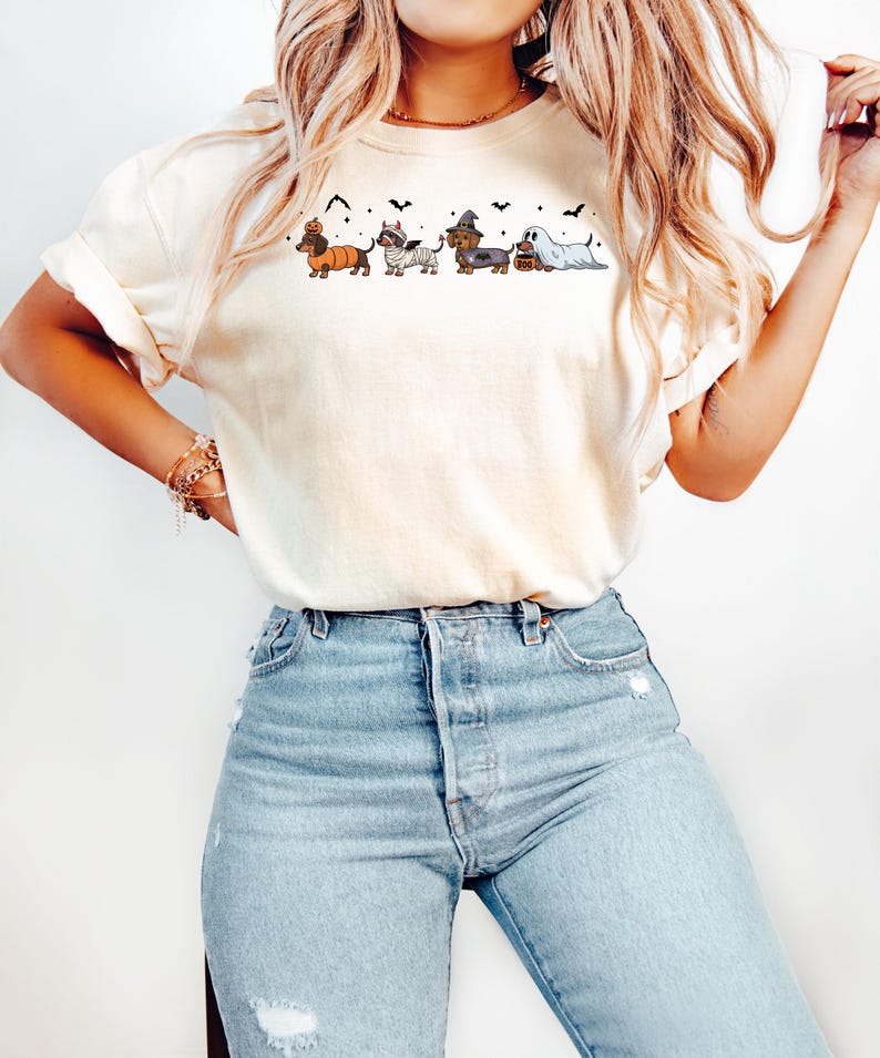 May include: Cream-colored t-shirt featuring a Halloween-themed dachshund design. The graphic includes dachshunds dressed as a pumpkin, mummy, witch, and ghost, with bats and a candy bucket. The shirt is paired with light blue jeans.
