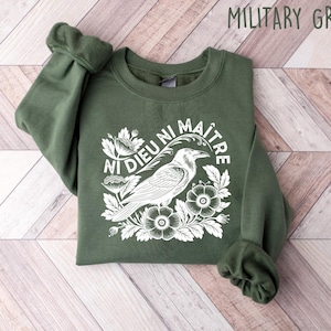 Op de afbeelding: Militairgroen sweatshirt met een witte afbeelding van een kraai, bloemen en de tekst "Ni Dieu Ni Ma&icirc;tre". De crewneck sweatshirt is opgevouwen op een houten oppervlak.