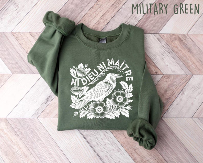 Op de afbeelding: Militairgroen sweatshirt met een witte afbeelding van een kraai, bloemen en de tekst "Ni Dieu Ni Ma&icirc;tre". De crewneck sweatshirt is opgevouwen op een houten oppervlak.