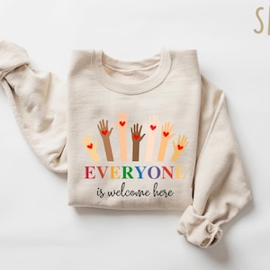May include: A sand-colored sweatshirt with a graphic of raised hands in various skin tones, each with a red heart. The text "EVERYONE is welcome here" is displayed in rainbow colors.