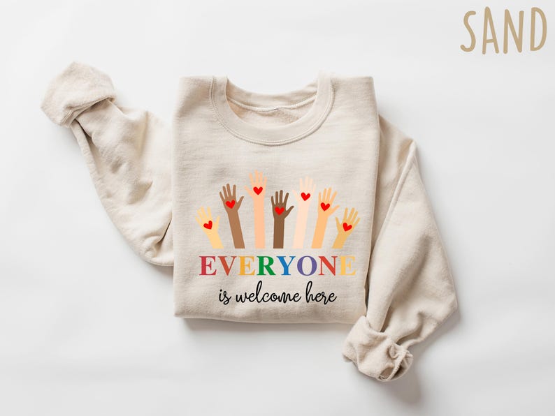 May include: A sand-colored sweatshirt with a graphic of raised hands in various skin tones, each with a red heart. The text "EVERYONE is welcome here" is displayed in rainbow colors.