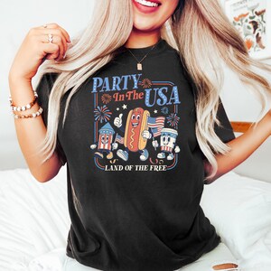 May include: Black t-shirt with a patriotic graphic. The design features cartoon illustrations of a firework, hot dog, and drink, along with the text "Party In The USA" and "Land of the Free."