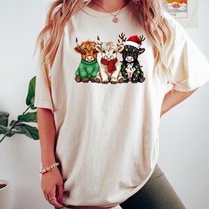 May include: Cream-colored t-shirt featuring a graphic of three Highland cows dressed for Christmas. One cow wears a green sweater, another a red scarf, and the third a Santa hat and Christmas lights. The shirt is a casual, festive apparel item.