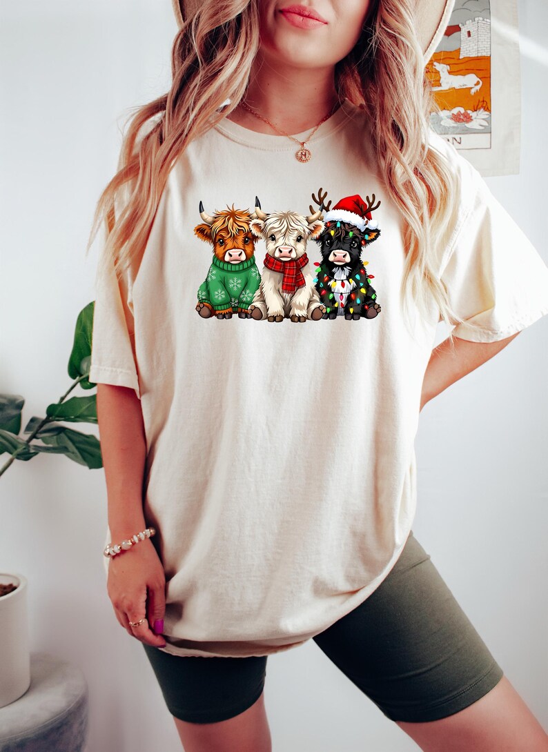May include: Cream-colored t-shirt featuring a graphic of three Highland cows dressed for Christmas. One cow wears a green sweater, another a red scarf, and the third a Santa hat and Christmas lights. The shirt is a casual, festive apparel item.