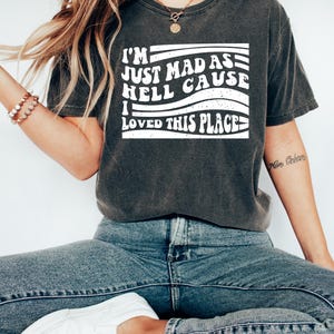 May include: Dark grey t-shirt featuring the white text "I'M JUST MAD AS HELL CAUSE I LOVED THIS PLACE". The shirt is worn with blue jeans and white trainers. The style is casual and relaxed.