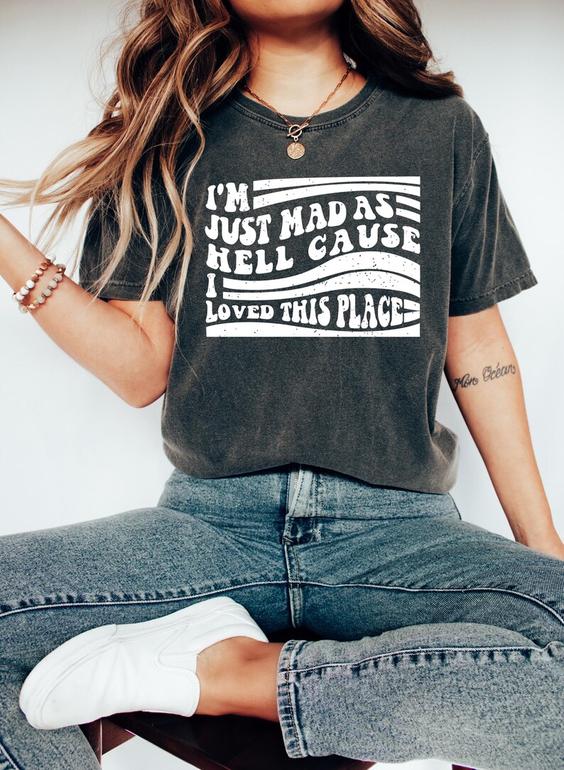 May include: Dark grey t-shirt featuring the white text "I'M JUST MAD AS HELL CAUSE I LOVED THIS PLACE". The shirt is worn with blue jeans and white trainers. The style is casual and relaxed.