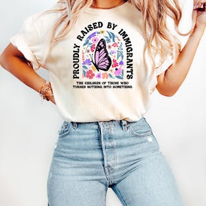 May include: Cream-colored t-shirt with the text "PROUDLY RAISED BY IMMIGRANTS" in a curved arch design. A pink butterfly is centered within the arch, surrounded by colorful flowers. The text "THE CHILDREN OF THOSE WHO TURNED NOTHING INTO SOMETHING" is below.
