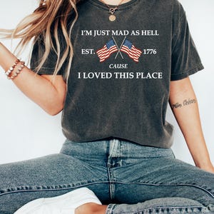 May include: Dark gray t-shirt with the text "I'M JUST MAD AS HELL CAUSE I LOVED THIS PLACE" and crossed American flags. The shirt also has "EST. 1776" printed on it. The person is wearing blue jeans and white shoes.