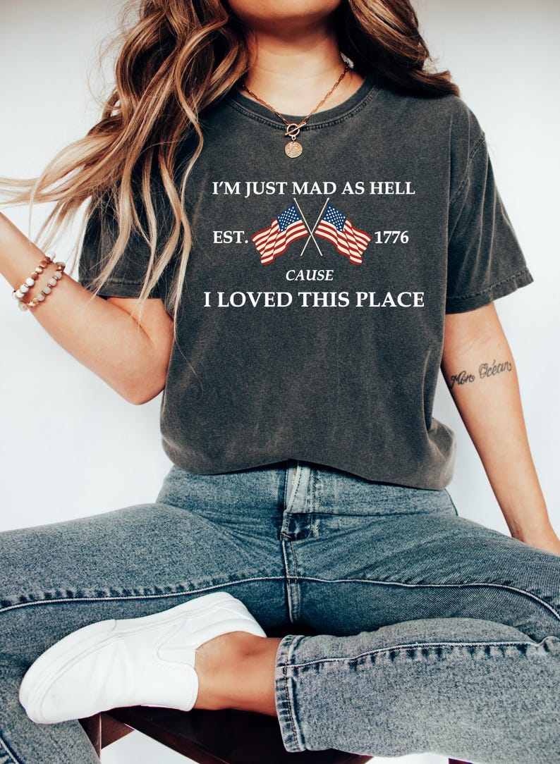 May include: Dark gray t-shirt with the text "I'M JUST MAD AS HELL CAUSE I LOVED THIS PLACE" and crossed American flags. The shirt also has "EST. 1776" printed on it. The person is wearing blue jeans and white shoes.
