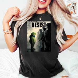 May include: Black t-shirt featuring a graphic of a green frog wearing a blue scarf, standing before a line of silhouetted figures. The word "RESIST" is printed above the frog. The shirt is worn by a person.