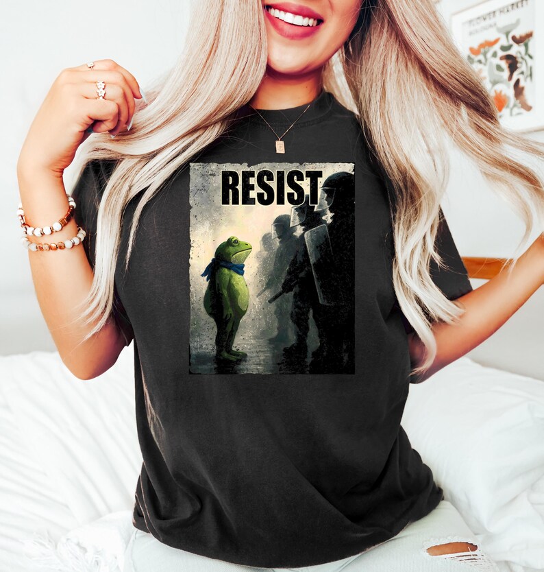 May include: Black t-shirt featuring a graphic of a green frog wearing a blue scarf, standing before a line of silhouetted figures. The word "RESIST" is printed above the frog. The shirt is worn by a person.