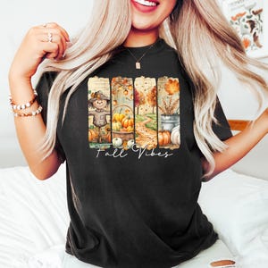 May include: Black t-shirt with a colorful fall-themed graphic. The design features a scarecrow, pumpkins, and a pathway, with the words "Fall Vibes" in a script font. The overall aesthetic is warm and autumnal.