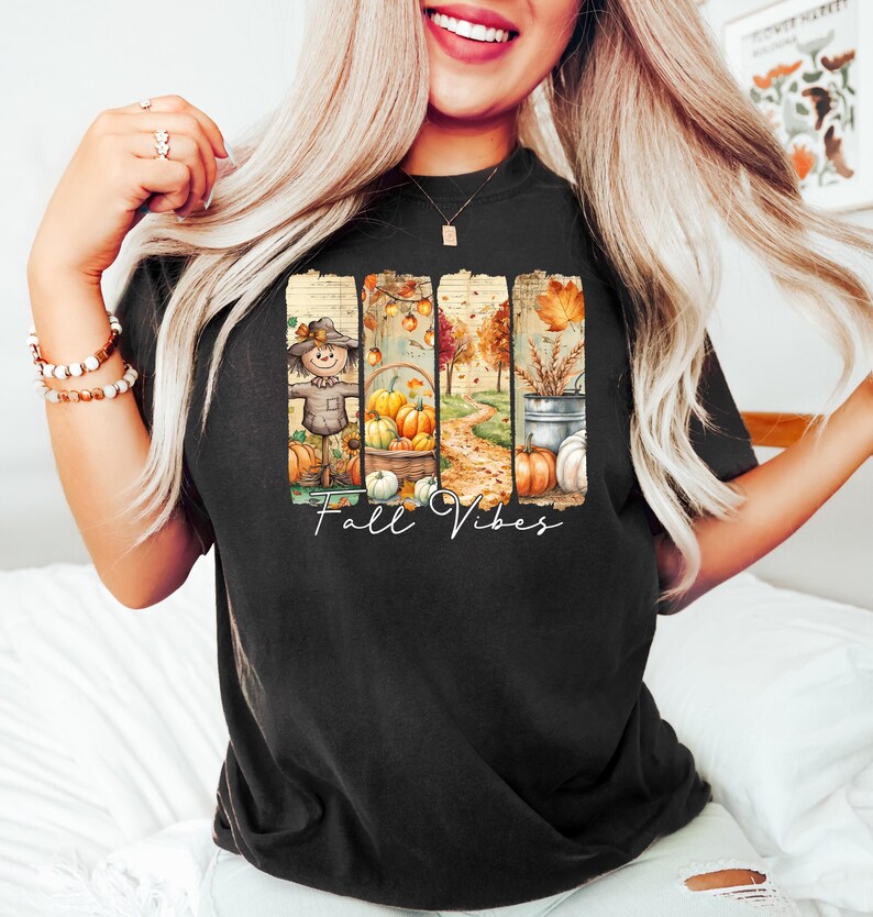 May include: Black t-shirt with a colorful fall-themed graphic. The design features a scarecrow, pumpkins, and a pathway, with the words "Fall Vibes" in a script font. The overall aesthetic is warm and autumnal.