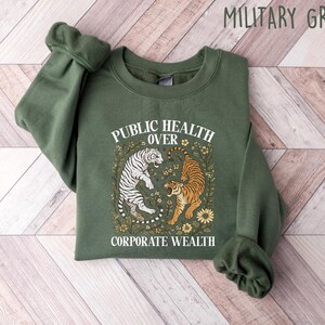 Puede incluir: Sudadera verde militar con el texto "PUBLIC HEALTH OVER CORPORATE WEALTH". El dise&ntilde;o presenta dos tigres rodeados de elementos florales. La sudadera est&aacute; hecha de un material suave.