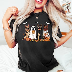 May include: Black t-shirt featuring a Halloween-themed graphic. The design includes four German Shepherd dogs dressed in costumes, such as a ghost, a cape, and bandages, with pumpkins and fall leaves. Bats are also included in the design.