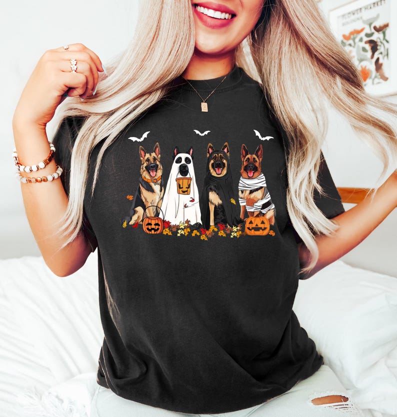 May include: Black t-shirt featuring a Halloween-themed graphic. The design includes four German Shepherd dogs dressed in costumes, such as a ghost, a cape, and bandages, with pumpkins and fall leaves. Bats are also included in the design.
