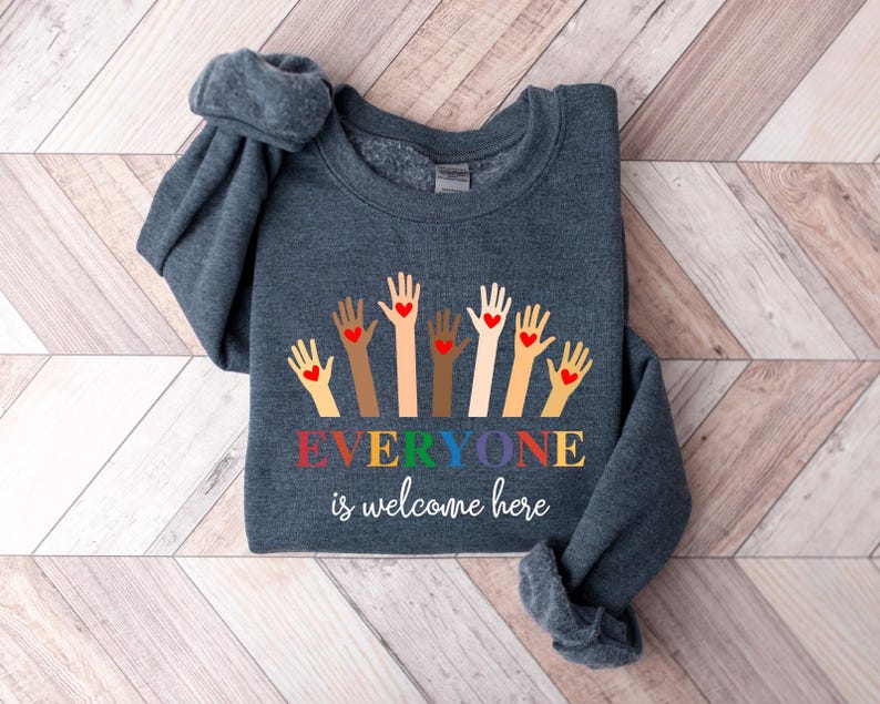 May include: A dark gray sweatshirt with the text "Everyone is welcome here." The design features hands of various skin tones, each with a red heart in the palm. The sweatshirt is folded on a light wood-grain surface.