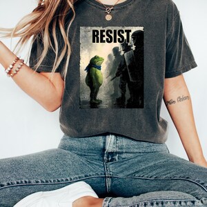 May include: Dark gray t-shirt featuring a graphic of a frog facing a line of riot police, with the word "RESIST" above. The frog wears a blue scarf. The shirt is a comfortable, casual style.