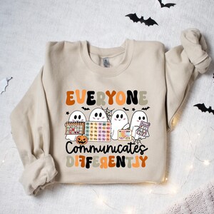 Może przedstawiać: Beżowa bluza z napisem "EVERYONE COMMUNICATES DIFFERENTLY" w kolorze pomarańczowym, czarnym i beżowym. Projekt zawiera cztery kresk&oacute;wkowe duszki trzymające r&oacute;żne przedmioty, w tym układ okresowy i dynię.
