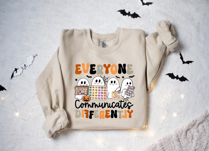 Może przedstawiać: Beżowa bluza z napisem "EVERYONE COMMUNICATES DIFFERENTLY" w kolorze pomarańczowym, czarnym i beżowym. Projekt zawiera cztery kresk&oacute;wkowe duszki trzymające r&oacute;żne przedmioty, w tym układ okresowy i dynię.