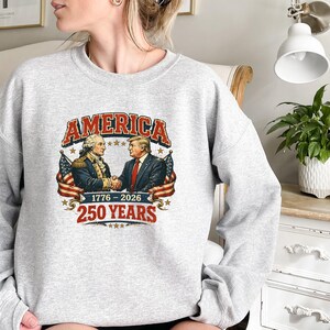 Puede incluir: Una sudadera gris claro con un dise&ntilde;o gr&aacute;fico con la palabra "AMERICA" en rojo y blanco. El dise&ntilde;o muestra a dos hombres d&aacute;ndose la mano, con banderas americanas y el texto "1776-2026 250 YEARS".