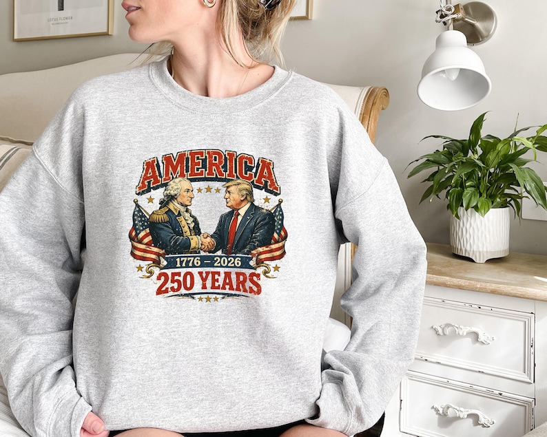 Puede incluir: Una sudadera gris claro con un dise&ntilde;o gr&aacute;fico con la palabra "AMERICA" en rojo y blanco. El dise&ntilde;o muestra a dos hombres d&aacute;ndose la mano, con banderas americanas y el texto "1776-2026 250 YEARS".