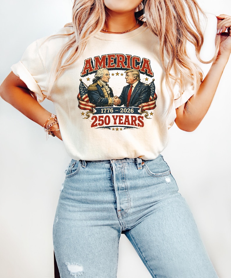 Puede incluir: Camiseta color crema con la palabra "AMERICA" en rojo desgastado. El dise&ntilde;o muestra a dos hombres estrech&aacute;ndose la mano, flanqueados por banderas americanas, con el texto "1776-2026" y "250 YEARS" debajo. La camiseta se combina con vaqueros azul claro.