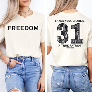 K&ouml;nnte beinhalten: Cremefarbenes T-Shirt mit dem Wort "FREEDOM" in gro&szlig;en, schwarzen Gro&szlig;buchstaben auf der Vorderseite. Auf der R&uuml;ckseite des Shirts steht der Text "THANK YOU, CHARLIE." &uuml;ber der Zahl "31", gef&uuml;llt mit Schwarz-Wei&szlig;-Bildern, und die Worte "A TRUE PATRIOT 1993-2025."