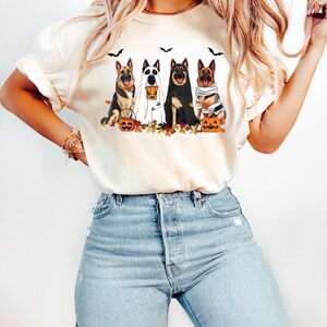 May include: Cream-colored t-shirt featuring a Halloween-themed graphic. The design showcases four German Shepherds dressed in costumes: a ghost, a vampire, a mummy, and a witch. Pumpkins, bats, and autumn leaves complete the festive scene.