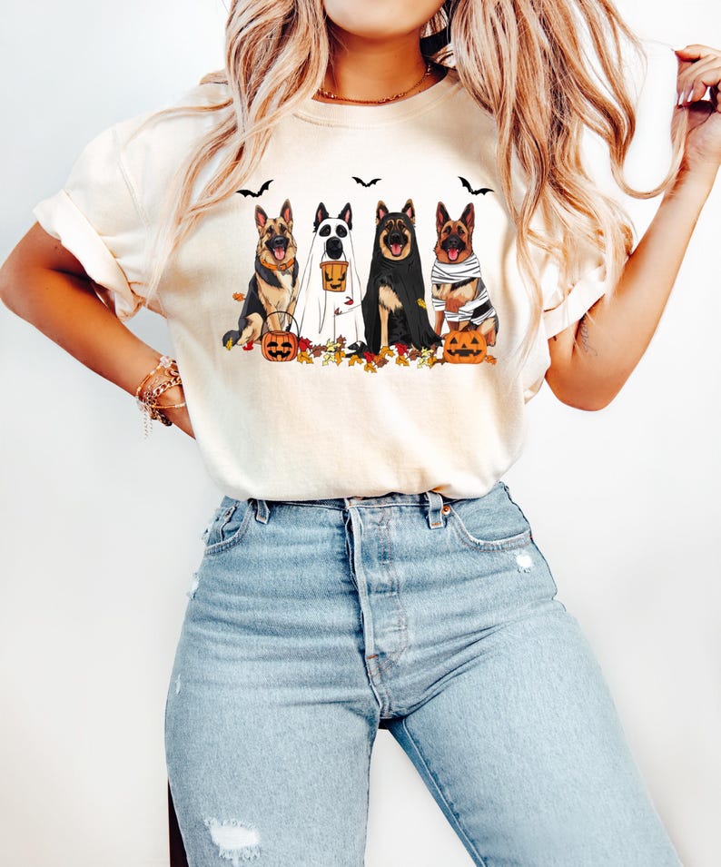 May include: Cream-colored t-shirt featuring a Halloween-themed graphic. The design showcases four German Shepherds dressed in costumes: a ghost, a vampire, a mummy, and a witch. Pumpkins, bats, and autumn leaves complete the festive scene.