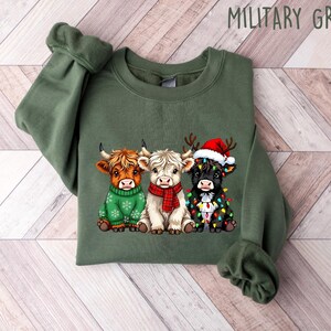 May include: Military green sweatshirt featuring a graphic of three Highland cows. One cow wears a green sweater, another a red scarf, and the third a Santa hat and Christmas lights. The text "MILITARY GREEN" is at the top.