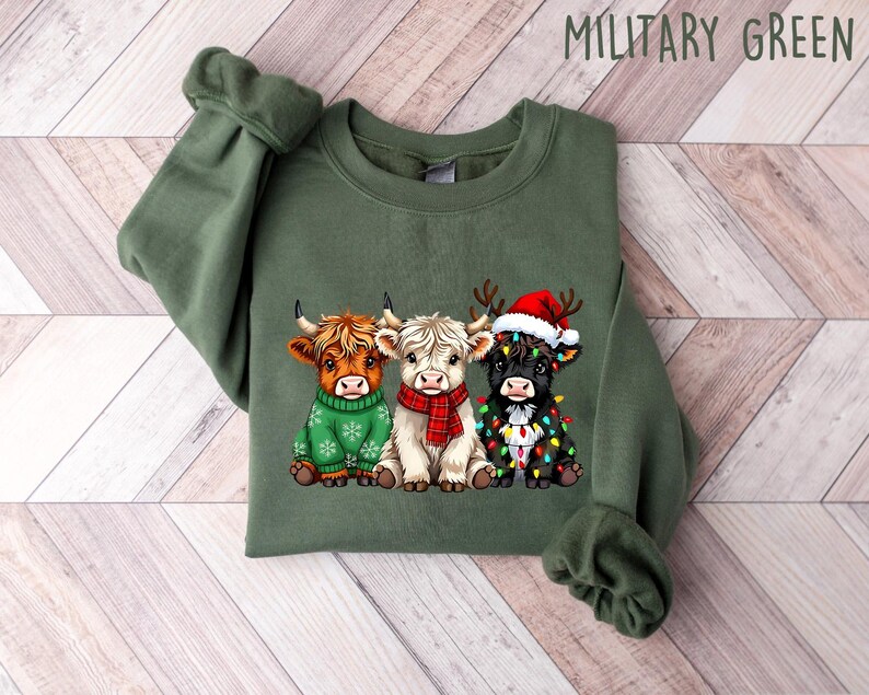 May include: Military green sweatshirt featuring a graphic of three Highland cows. One cow wears a green sweater, another a red scarf, and the third a Santa hat and Christmas lights. The text "MILITARY GREEN" is at the top.