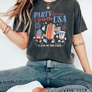 May include: Dark gray t-shirt with a retro graphic that says "PARTY In The USA" with cartoon characters of a firework, hot dog, and coffee cup holding American flags. The text "LAND OF THE FREE" is below the graphic.