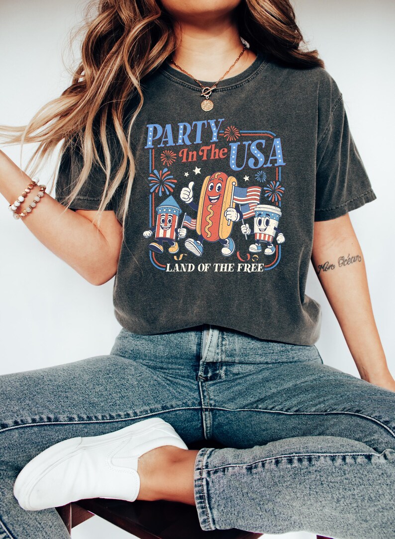 May include: Dark gray t-shirt with a retro graphic that says "PARTY In The USA" with cartoon characters of a firework, hot dog, and coffee cup holding American flags. The text "LAND OF THE FREE" is below the graphic.
