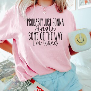 Op de afbeelding: Een lichtroze t-shirt met de zwarte tekst "PROBABLY JUST GONNA jingle SOME OF THE WAY I'm tired". Een witte mok met een gele smiley wordt vastgehouden. De persoon draagt lichtblauwe shorts.