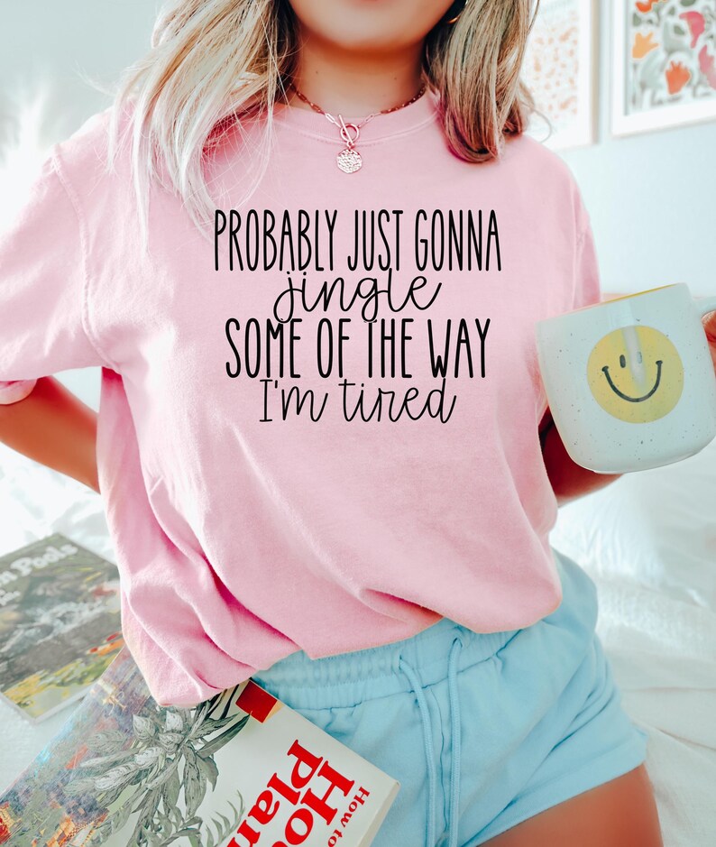 Op de afbeelding: Een lichtroze t-shirt met de zwarte tekst "PROBABLY JUST GONNA jingle SOME OF THE WAY I'm tired". Een witte mok met een gele smiley wordt vastgehouden. De persoon draagt lichtblauwe shorts.