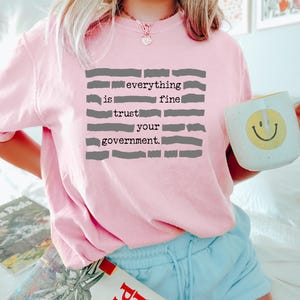 Puede incluir: Sudadera rosa claro con el texto gris "everything is fine trust your government". Se sostiene una taza blanca con una carita sonriente amarilla. La persona lleva pantalones cortos azul claro.