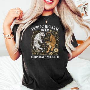 Puede incluir: Camiseta negra con el texto "PUBLIC HEALTH OVER CORPORATE WEALTH" y un gr&aacute;fico de dos tigres. Un tigre es blanco con rayas negras, el otro naranja con rayas negras, rodeados de elementos florales.