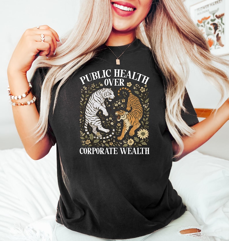 Puede incluir: Camiseta negra con el texto "PUBLIC HEALTH OVER CORPORATE WEALTH" y un gr&aacute;fico de dos tigres. Un tigre es blanco con rayas negras, el otro naranja con rayas negras, rodeados de elementos florales.